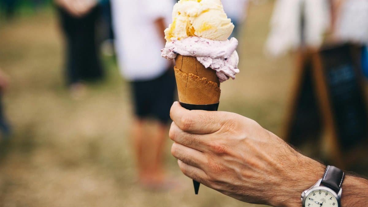Hand holding an ice cream cone with multiple flavors outdoors on a sunny day.