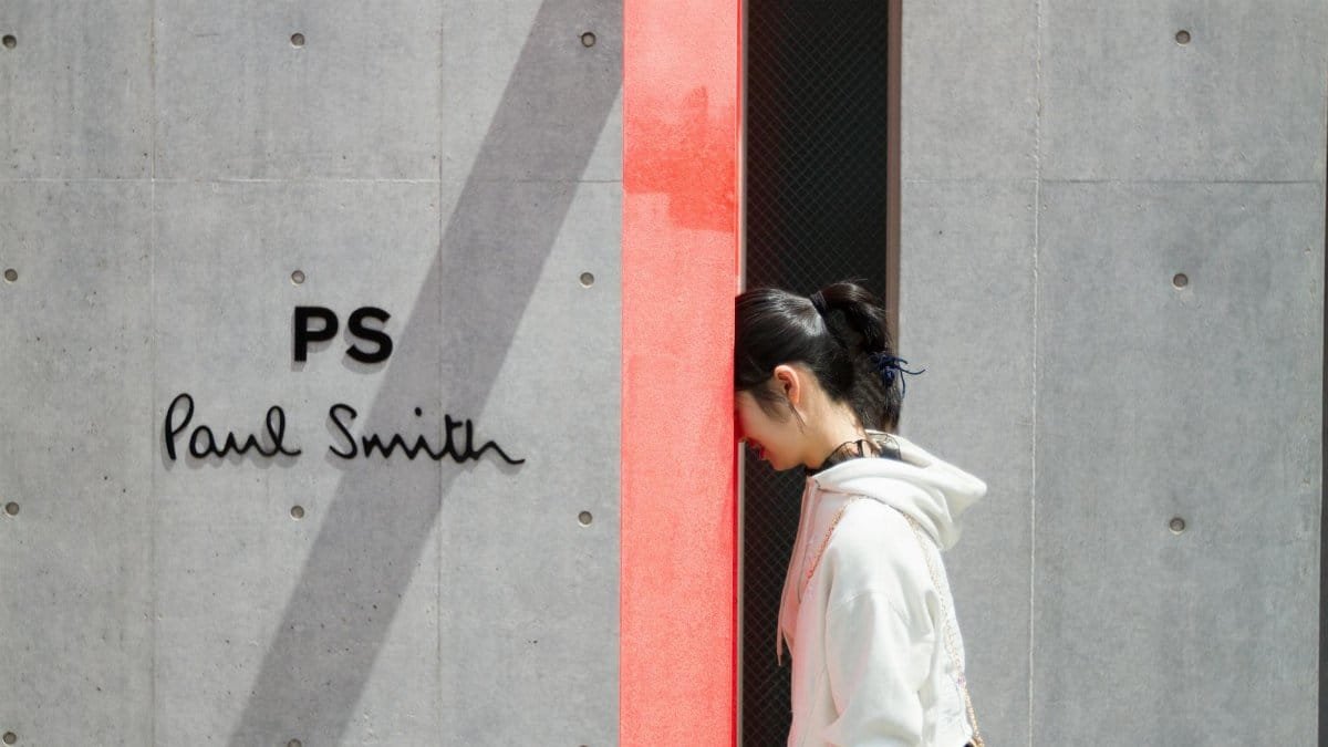 A woman in a white hoodie walks past a minimalist Paul Smith store sign and wall.
