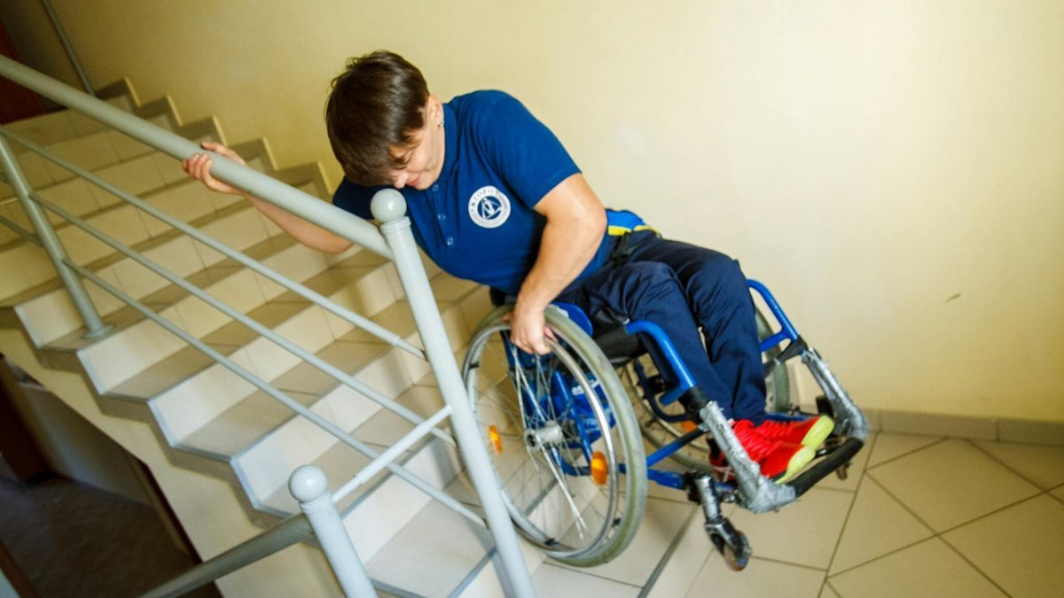 A person in a wheelchair navigating stairs with a handrail indoors, representing accessibility challenges.