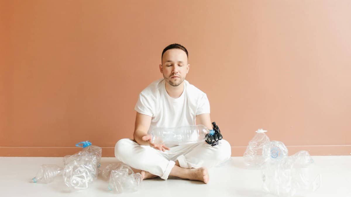 Man with prosthetic hand meditates indoors among plastic bottles, promoting environmental awareness.