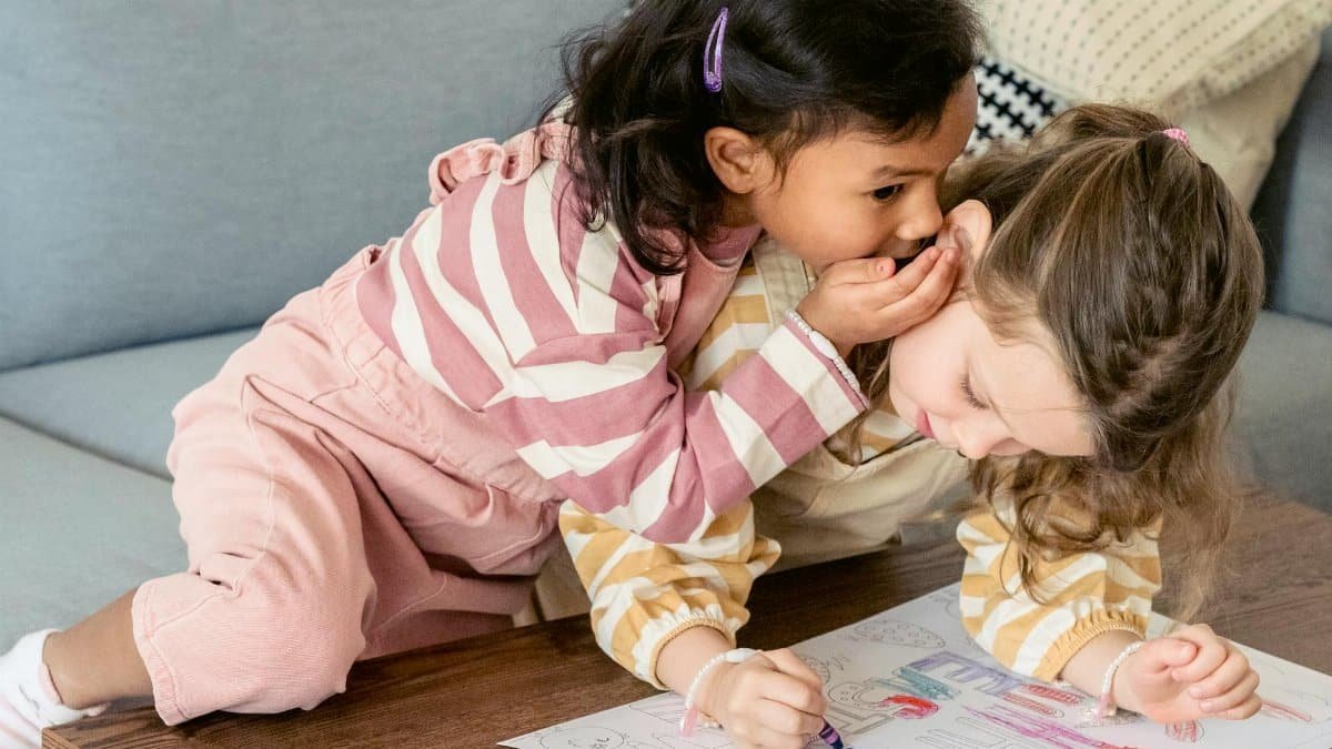 Attentive Hispanic child whispering in ear of girlfriend coloring paper drawing with crayon at table in house