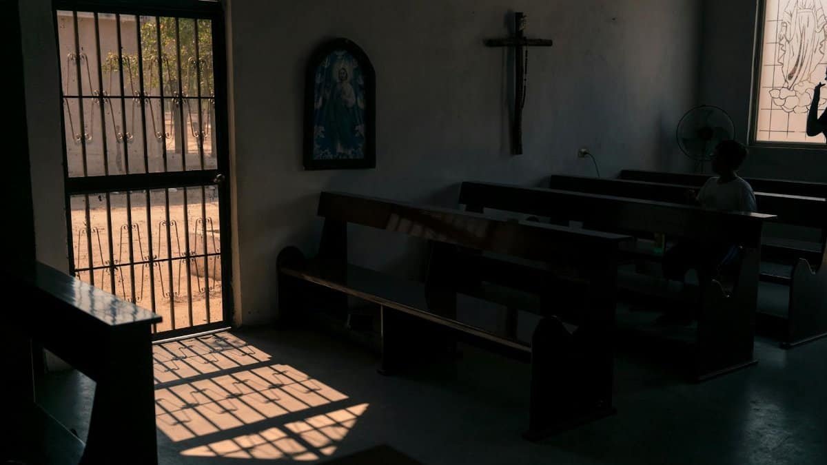 Dark chapel with cross on wall and wooden benches located in city with metal gate casting shadow on floor on sunny day