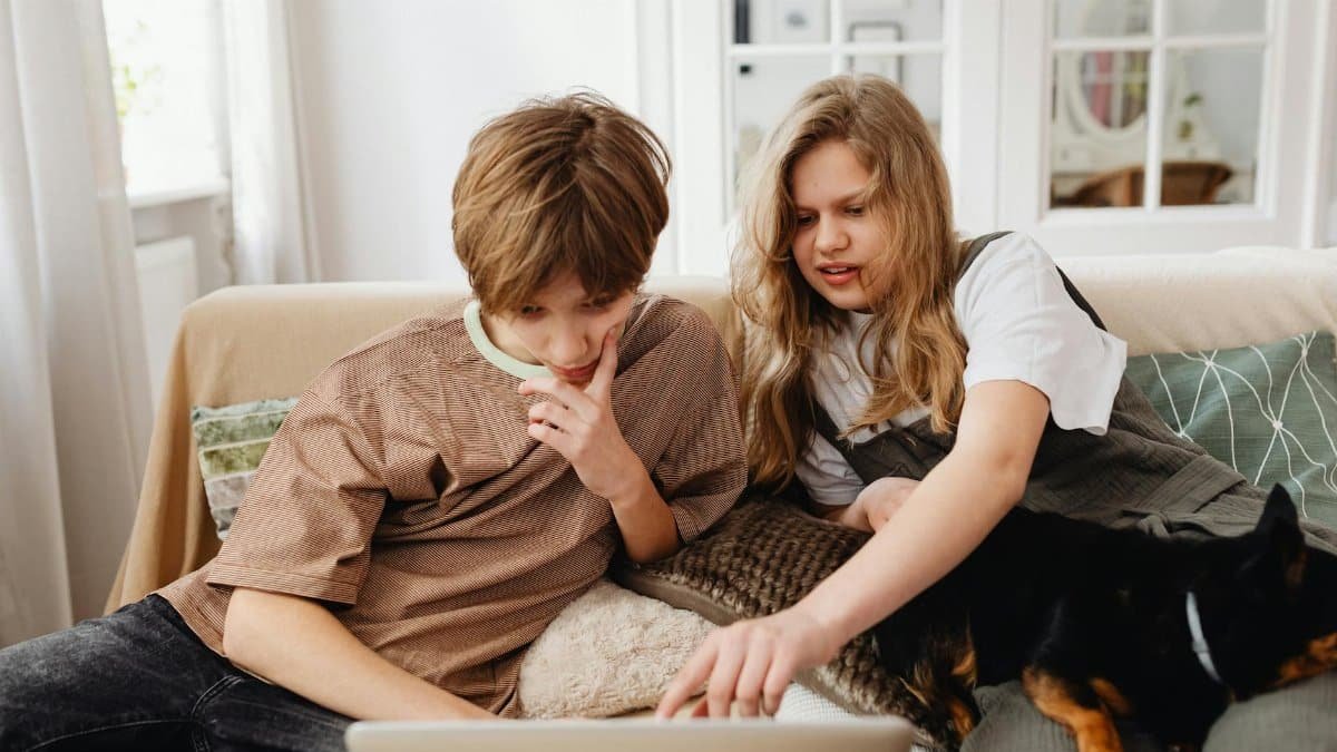 Two siblings with their dog enjoy quality time on a sofa using a laptop together.