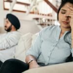 A young couple sitting on a couch indoors, showing signs of stress and frustration.