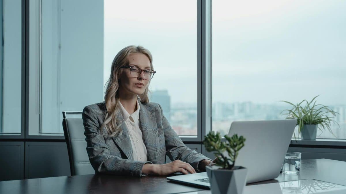 A confident businesswoman in a grey suit working on a laptop in a modern office setting.