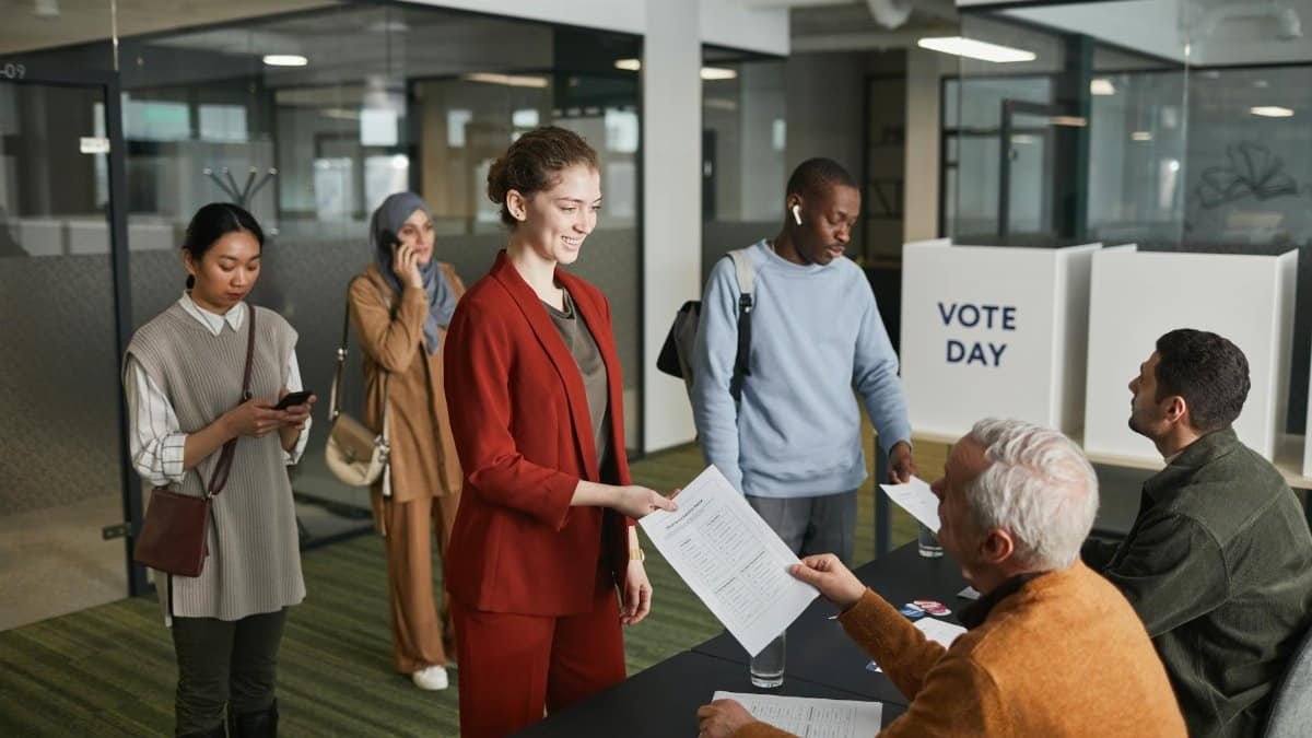 A diverse group of adults participating in an indoor voting process, engaging with election officials.