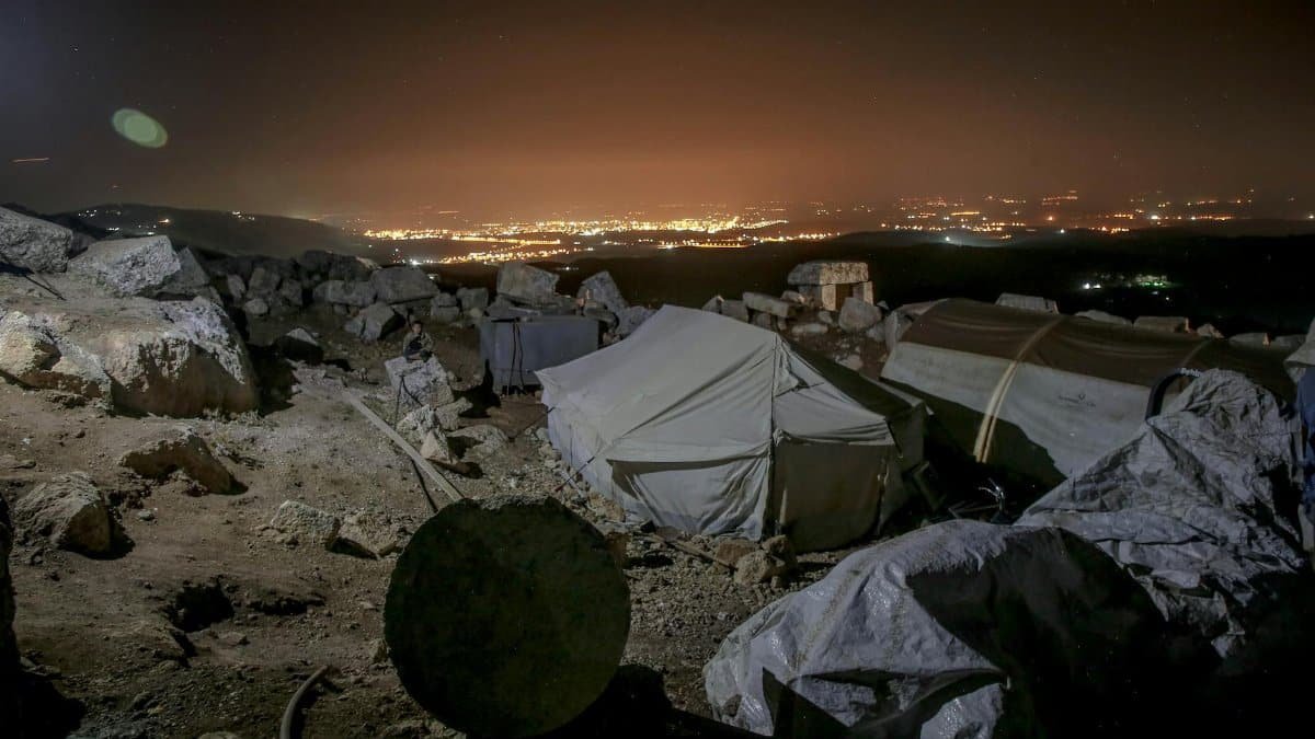 Refugee tents amidst rocky terrain with city lights at night in Idlib, Syria.