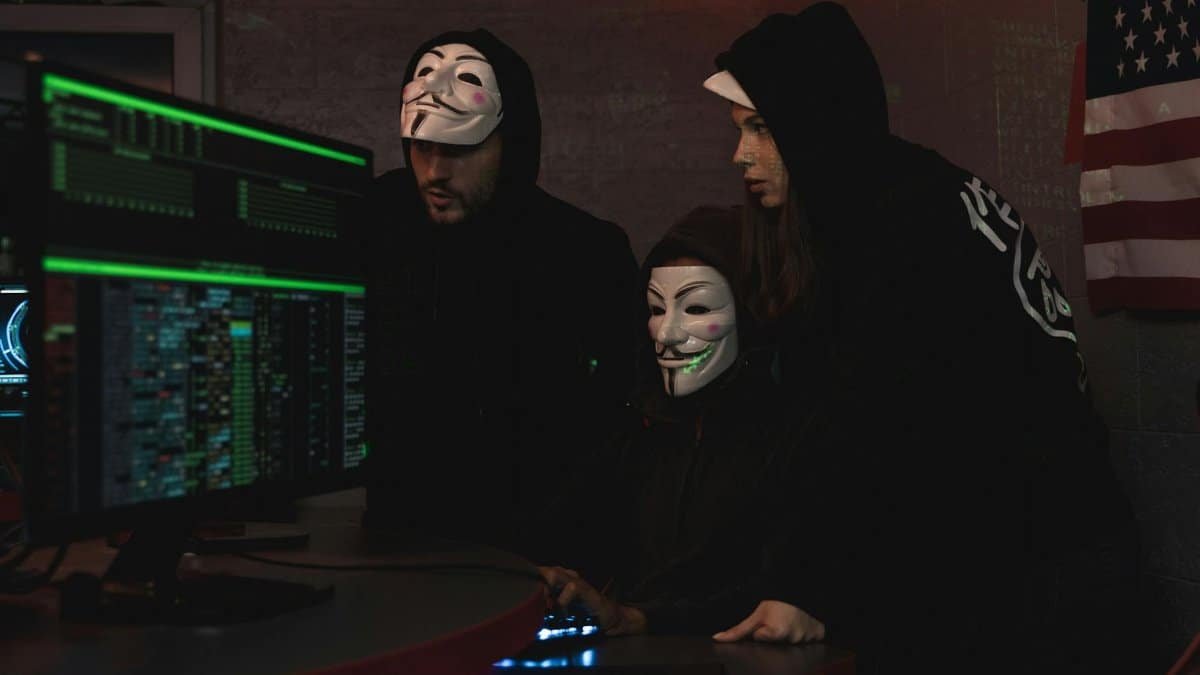 Masked hackers in hoodies working on a system in a dimly lit room, representing cyber security threats.