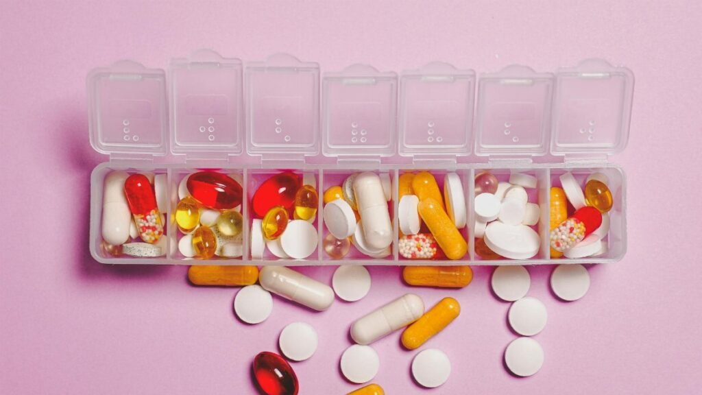 A variety of pills in a weekly pill organizer on a pink background.