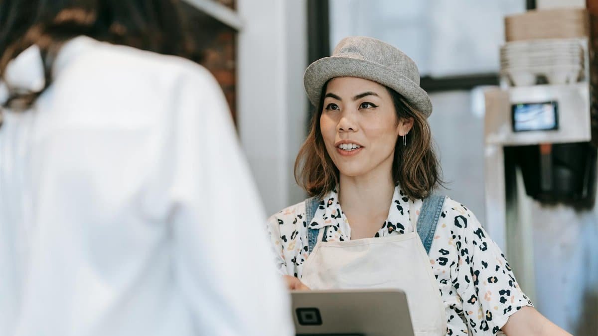 A cheerful barista in a hat takes an order from a customer at a cozy coffee shop counter.