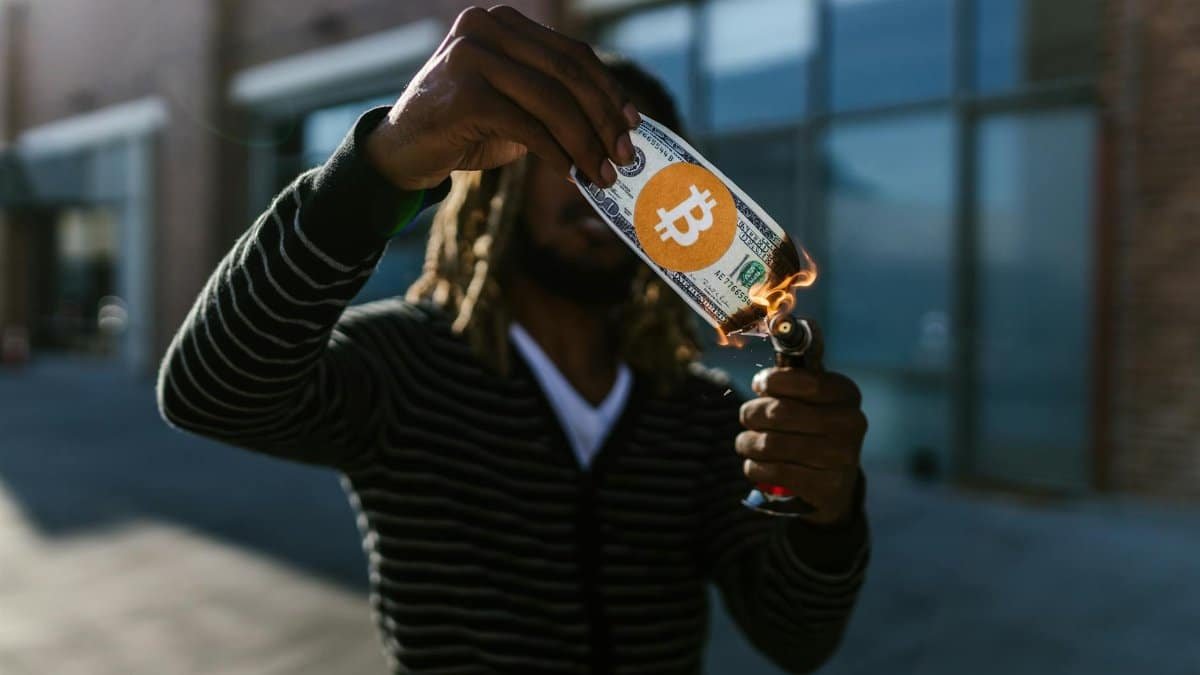 A man burns a fake dollar with a Bitcoin symbol outdoors, symbolizing cryptocurrency risks.
