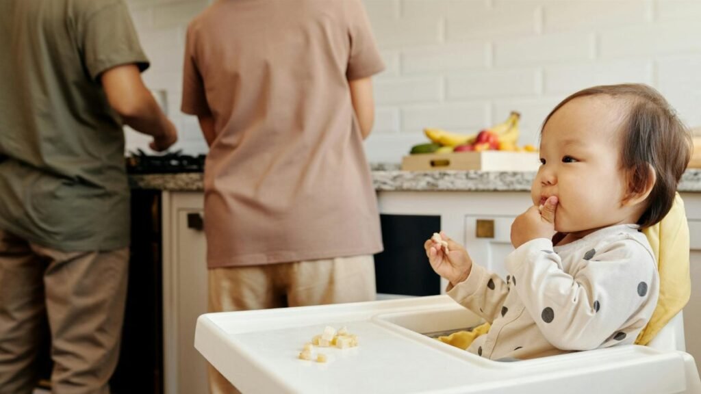 Cute toddler sits in a high chair enjoying a meal, while parents cook in the background of a modern kitchen.