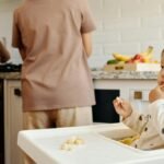 Cute toddler sits in a high chair enjoying a meal, while parents cook in the background of a modern kitchen.