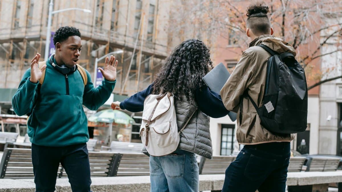 Back view of faceless student with backpack pushing away diverse classmates while standing in city park