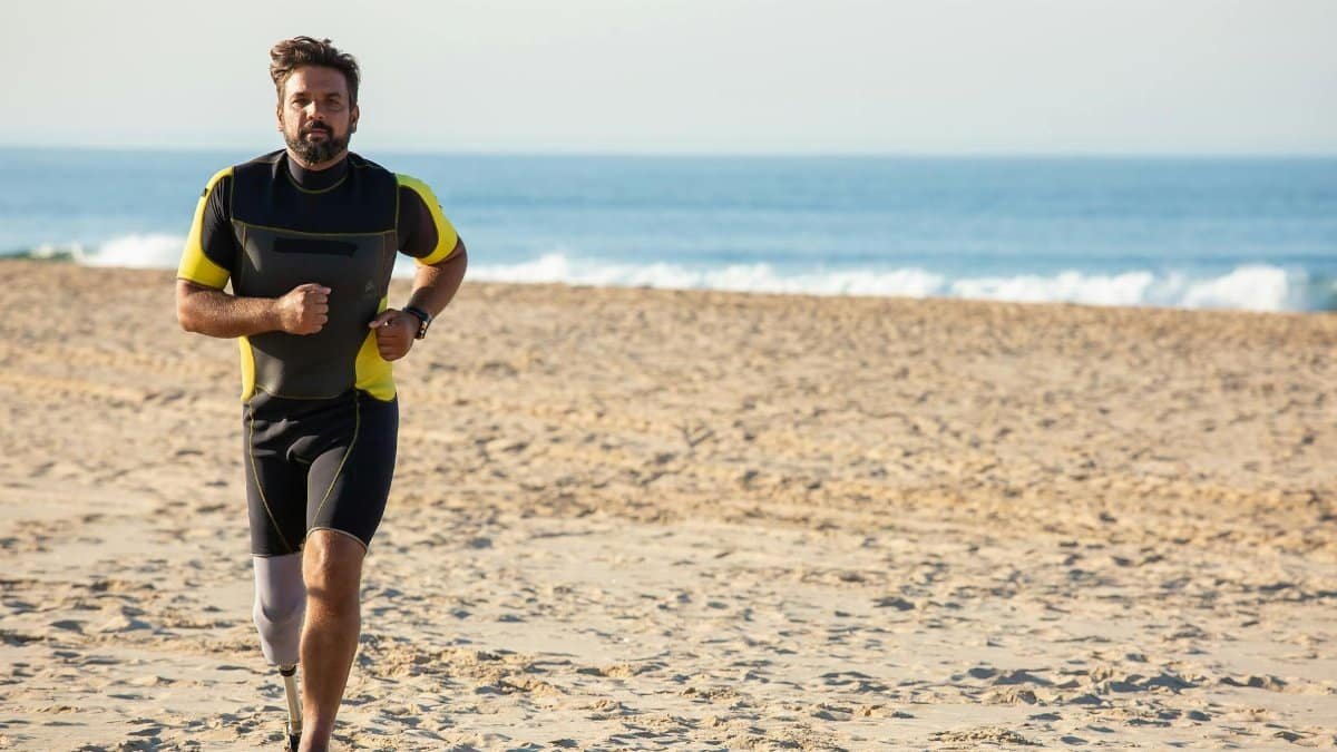 Full body energetic ethnic male with leg prosthesis in activewear jogging on sunny sandy seashore against calm sea and looking at camera