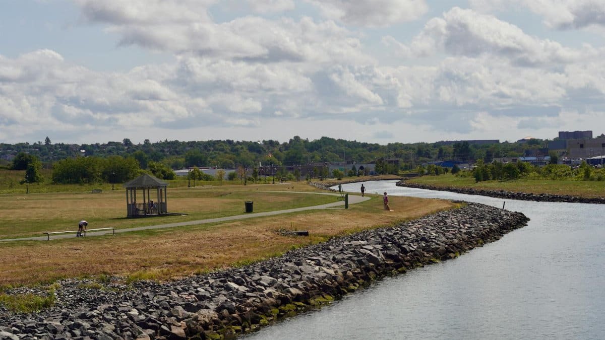 A serene riverside landscape featuring a gazebo, walking path, and distant hills under a cloudy sky.