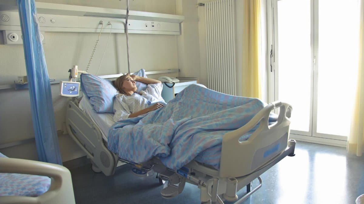 A woman lying in a hospital bed in a bright, softly lit room, appearing to rest or recover.