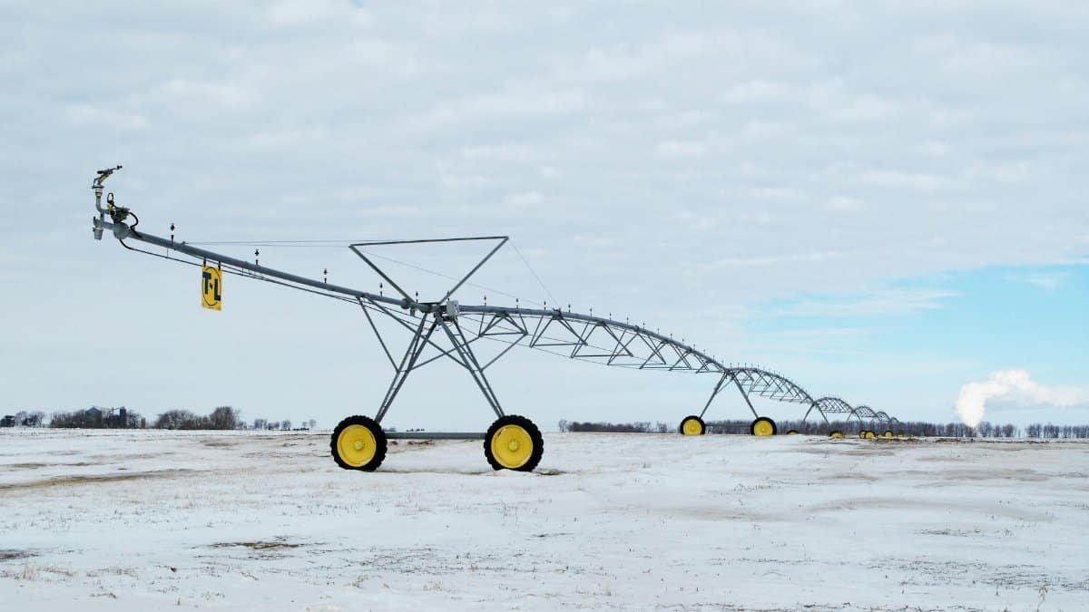 A snow-covered agricultural field with a large metal irrigation sprinkler system under a cloudy sky.