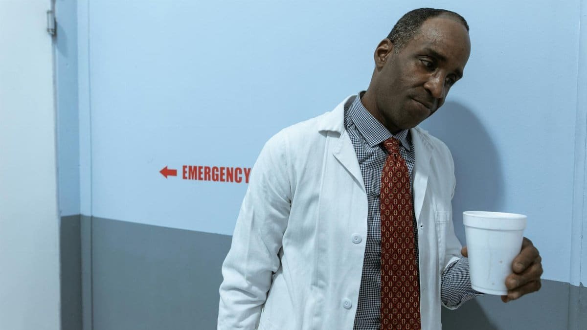 A healthcare worker in a lab coat holding a cup in a hospital corridor by the emergency section.