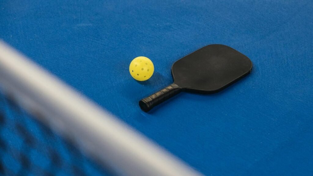A pickleball paddle and ball resting on a blue court surface with a net in focus.