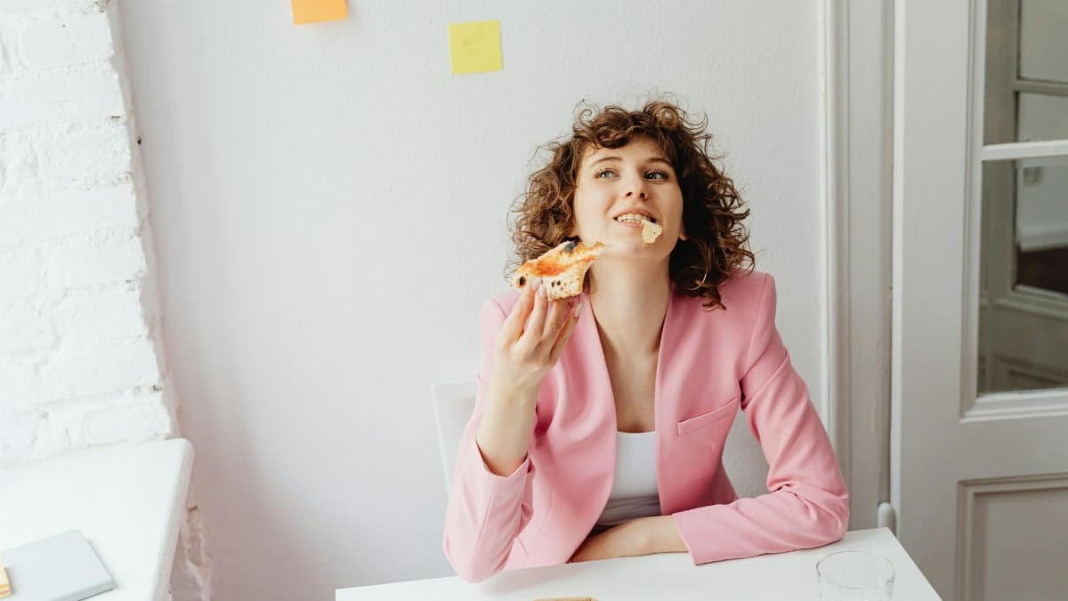 A cheerful woman in a pink blazer takes a moment to enjoy pizza while working from home.