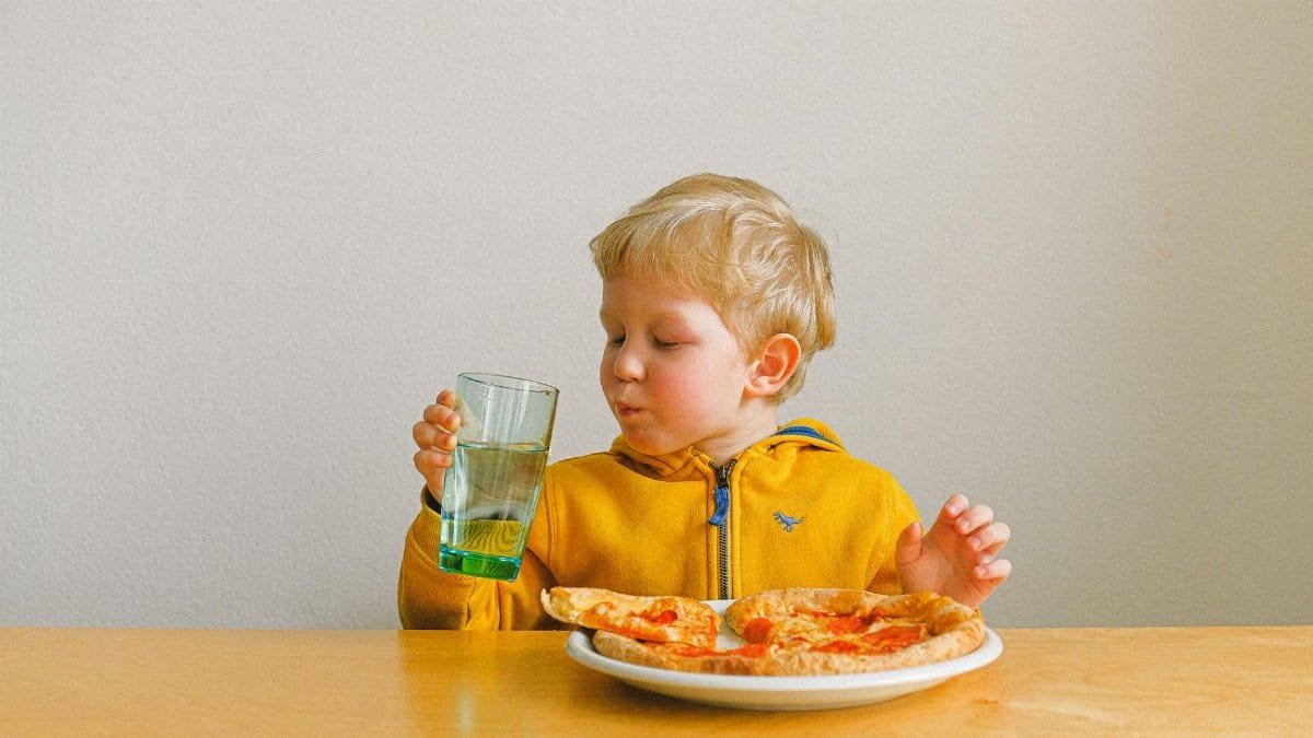 A cute blond child happily drinks while enjoying a pizza meal indoors.