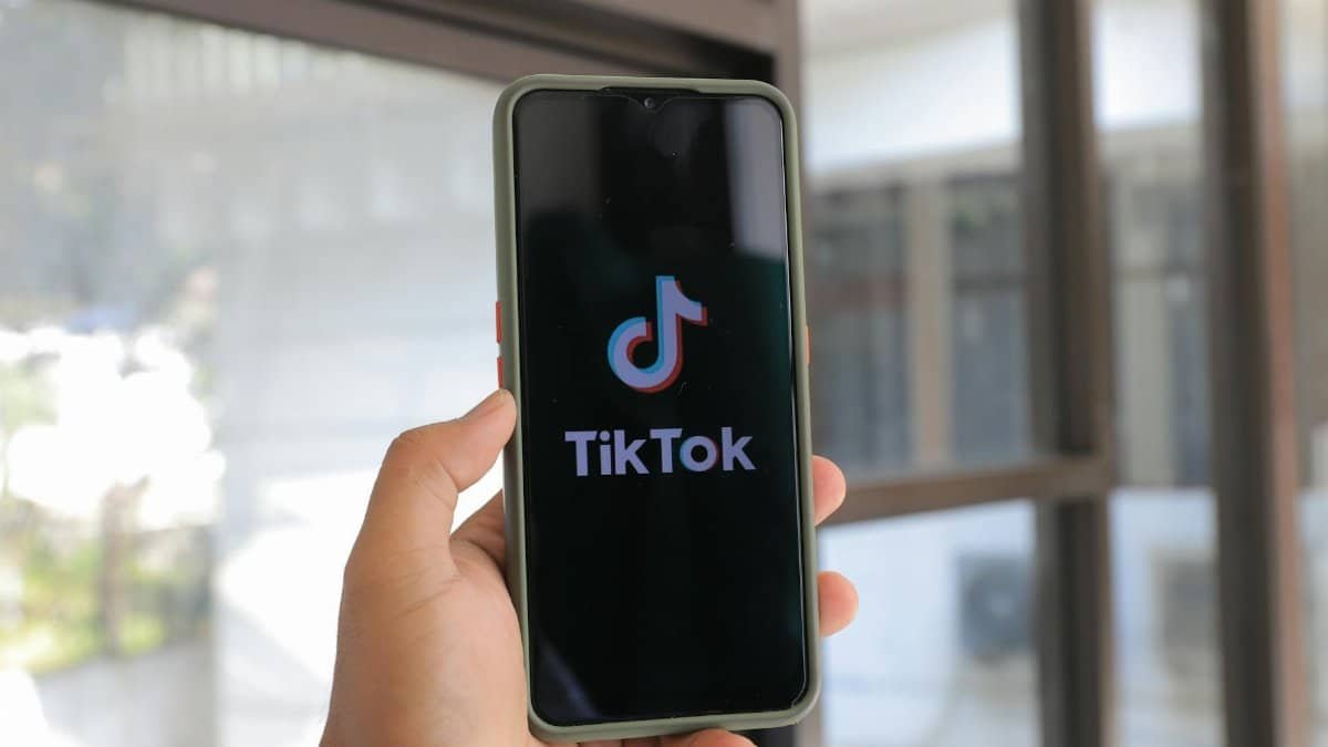 A hand holds a smartphone displaying the TikTok app in an indoor setting.