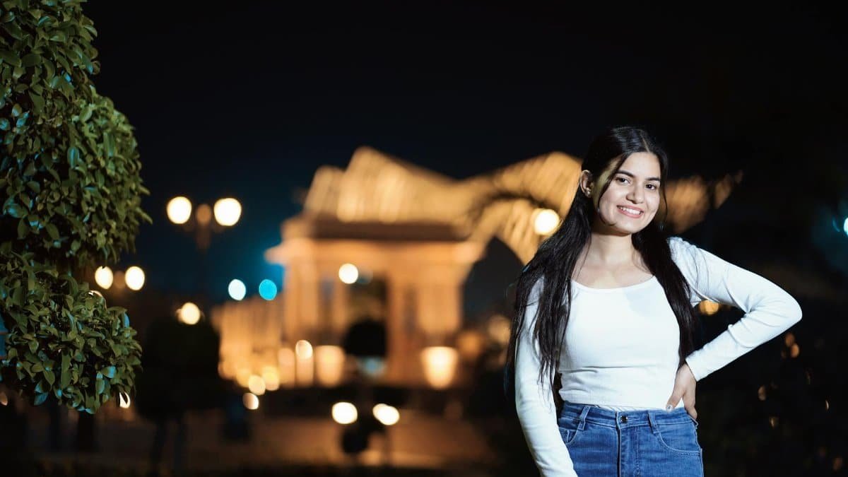 A young woman poses confidently outdoors at night with a lit building in the background.