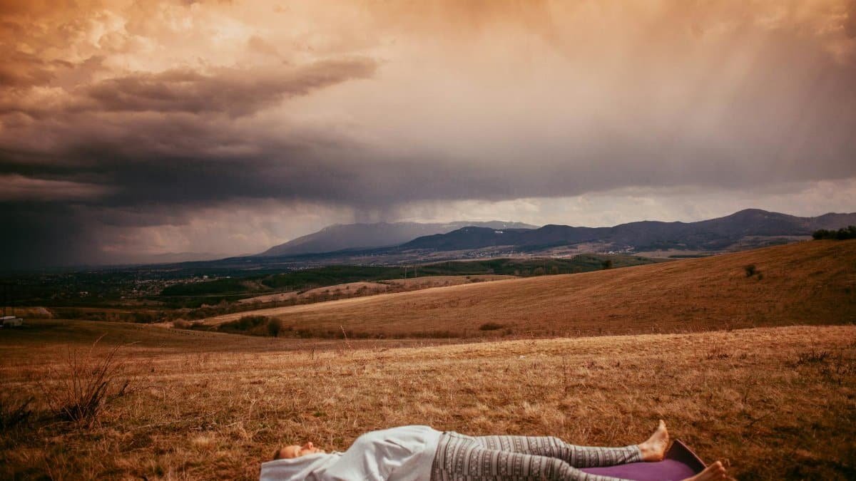 Person meditating on a yoga mat in a serene countryside setting with moody clouds.