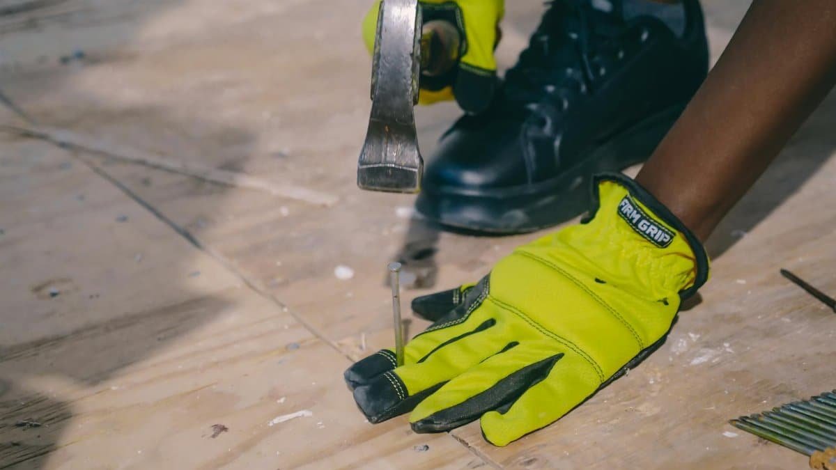 A worker wearing bright gloves uses a hammer on a plywood surface, emphasizing safety and construction.