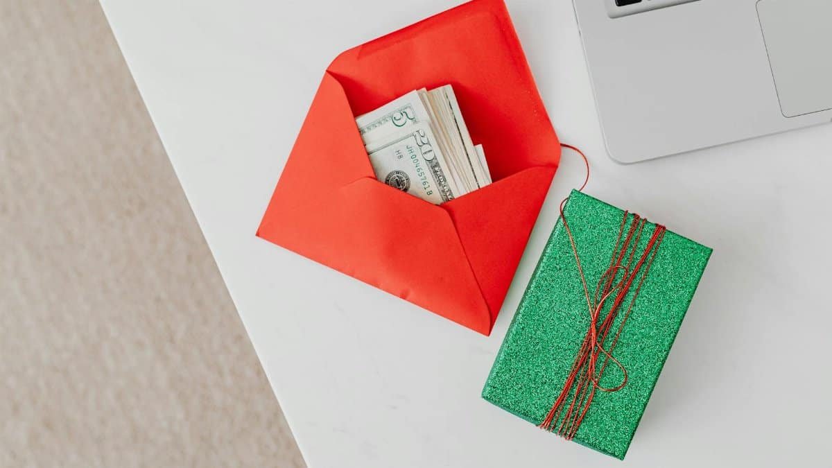 A green gift box and red envelope with cash on a desk beside a laptop, emphasizing holiday or financial themes.