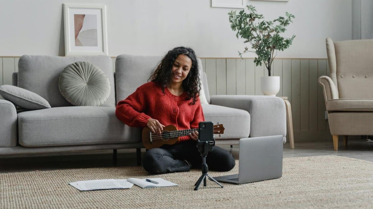 Smiling woman playing ukulele in cozy room with laptop and recording setup.