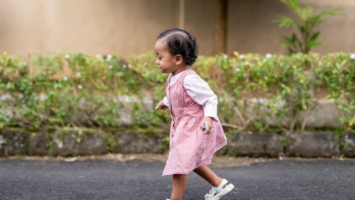 Cute toddler in a pink dress walking on a pathway with green foliage background, showing early steps.
