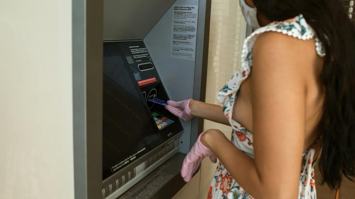 Woman with mask and gloves withdrawing cash from ATM. Safety measures during pandemic.