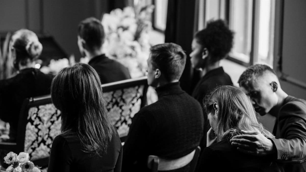A somber grayscale image of mourners sitting indoors during a funeral, conveying loss and sympathy.