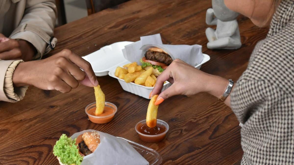 Friends sharing a meal of hamburgers and fries, dipping in sauces at a wooden table.