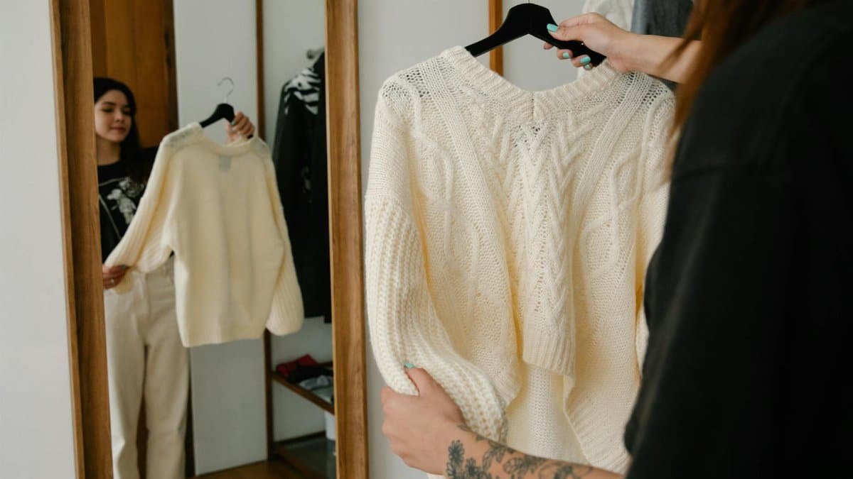 Woman holding and trying on a knitted sweater while observing her reflection in a mirror, showcasing fashion styling.