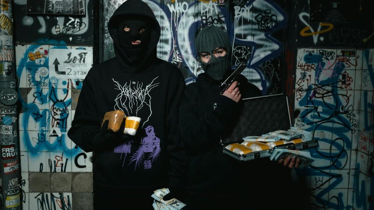 Two masked individuals holding cash, gun, and briefcase in a graffiti-covered alley.