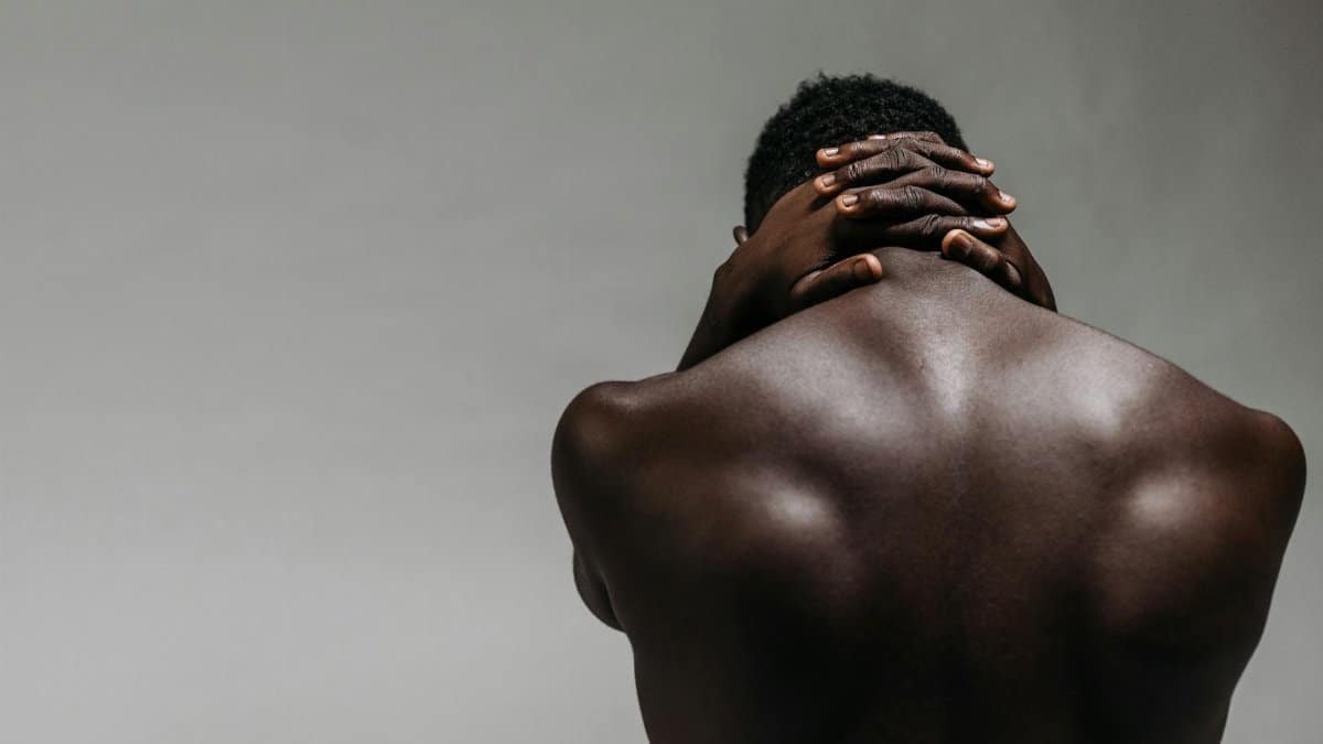 Back view of a brawny shirtless man with hands clasped on neck against a gray background.