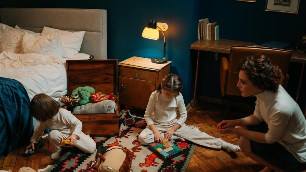 Children engaging with toys in a cozy bedroom, supervised by a nanny. Warm indoor scene.