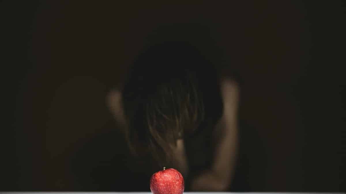 A solitary red apple on a table with a blurred, thoughtful figure in the background.