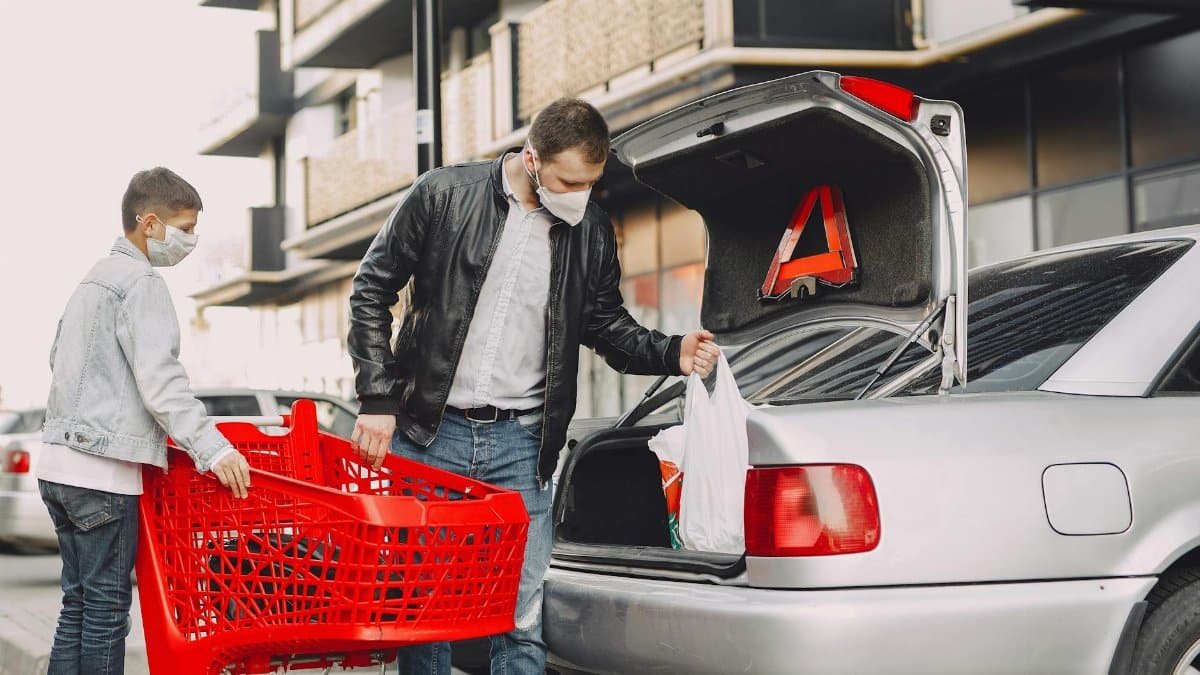 Father and son wearing masks unload groceries into car trunk during pandemic shopping trip.
