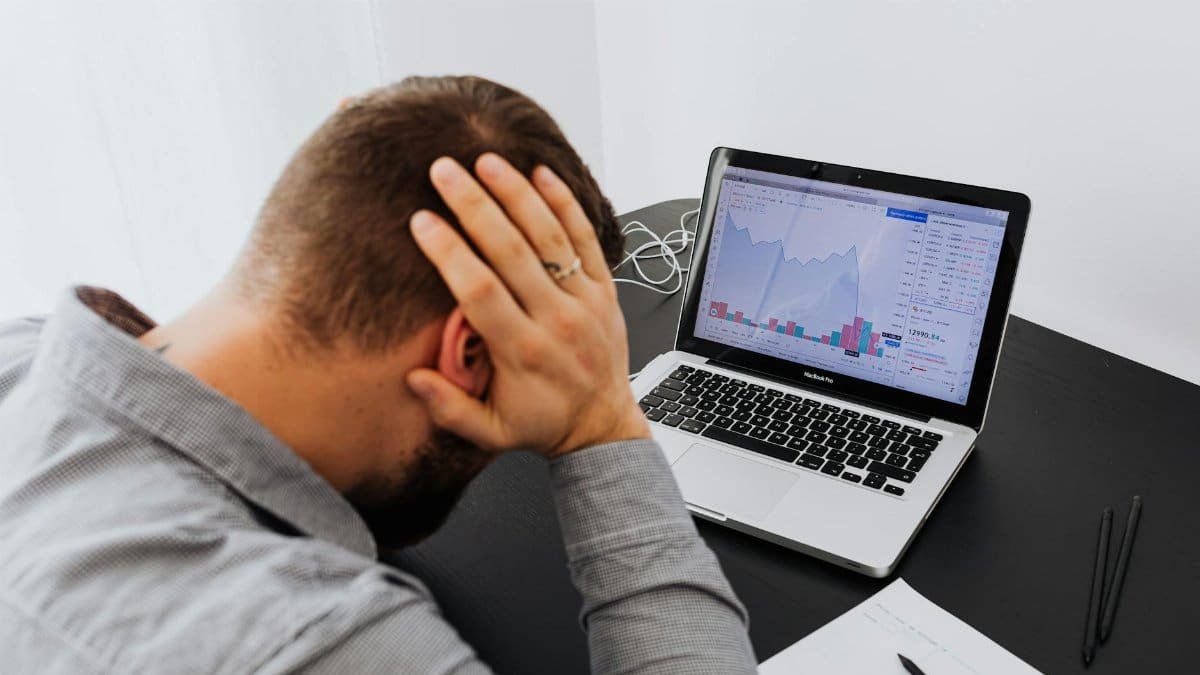 Stressed man at desk looking at declining stock charts on laptop, indicating financial loss.
