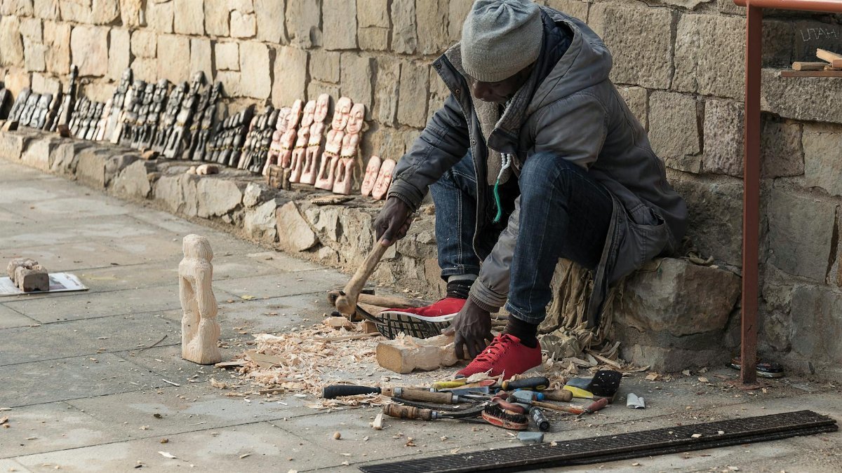 A street artist skillfully carves wooden sculptures on a city sidewalk, showcasing intricate craftsmanship.