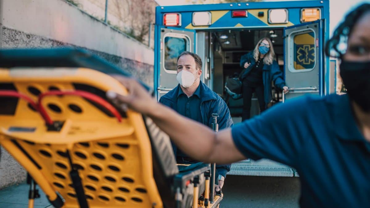 Paramedics wearing masks load a stretcher into an ambulance during a daytime emergency response.