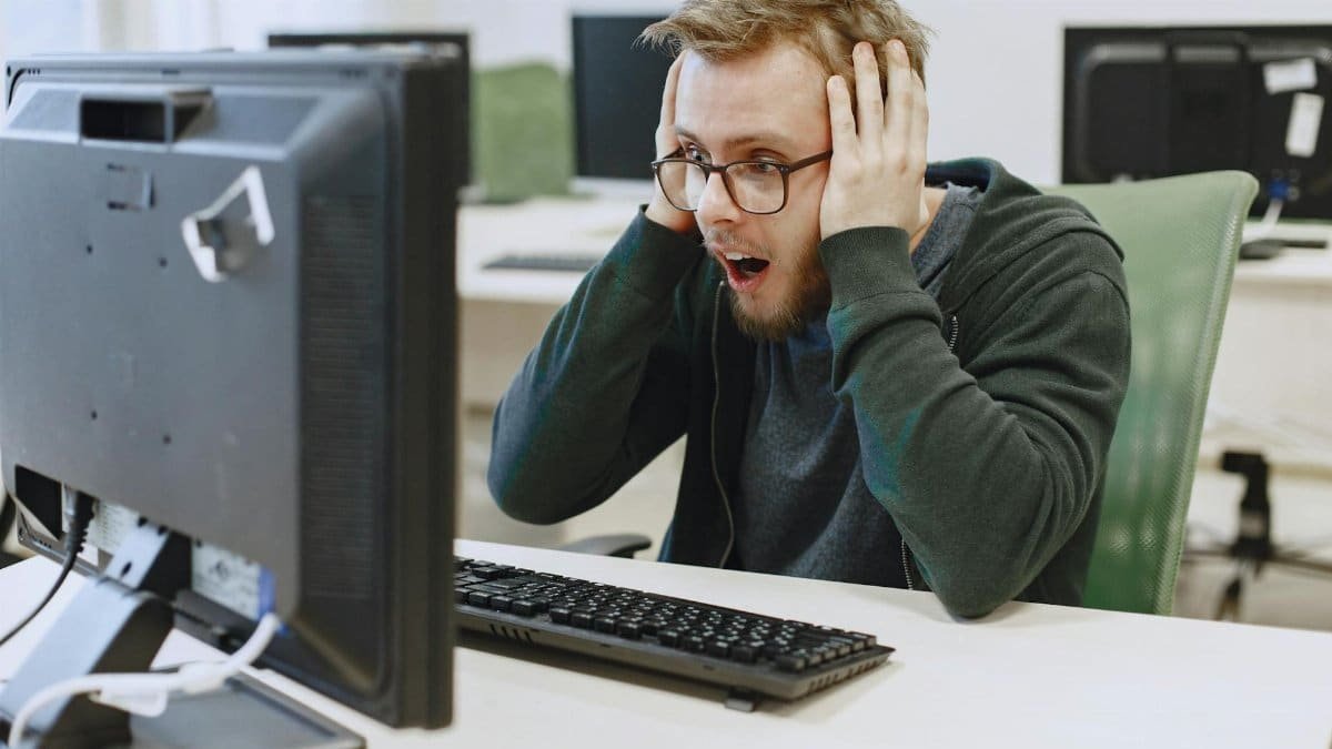 Surprised man in hoodie reacting to shocking news on computer monitor while sitting in a modern office.