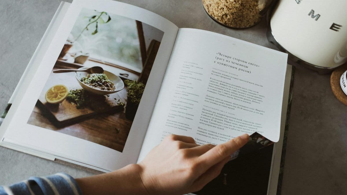 A person turns the page of a recipe book on a kitchen counter, surrounded by cooking ingredients.