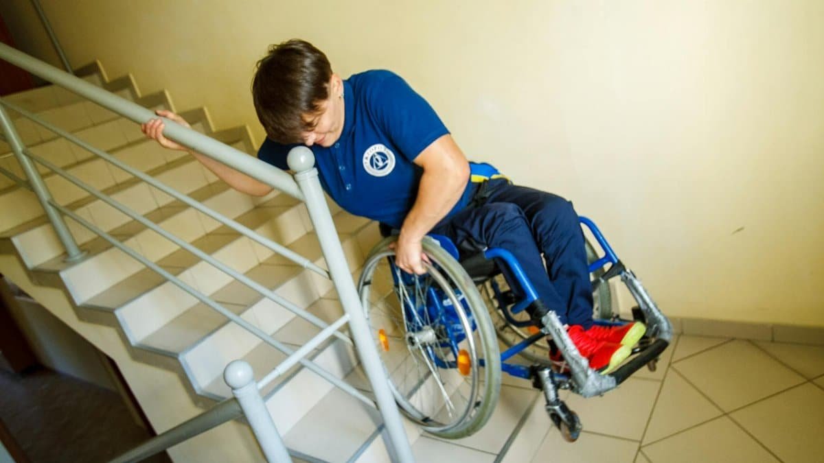 A person in a wheelchair navigating stairs with a handrail indoors, representing accessibility challenges.