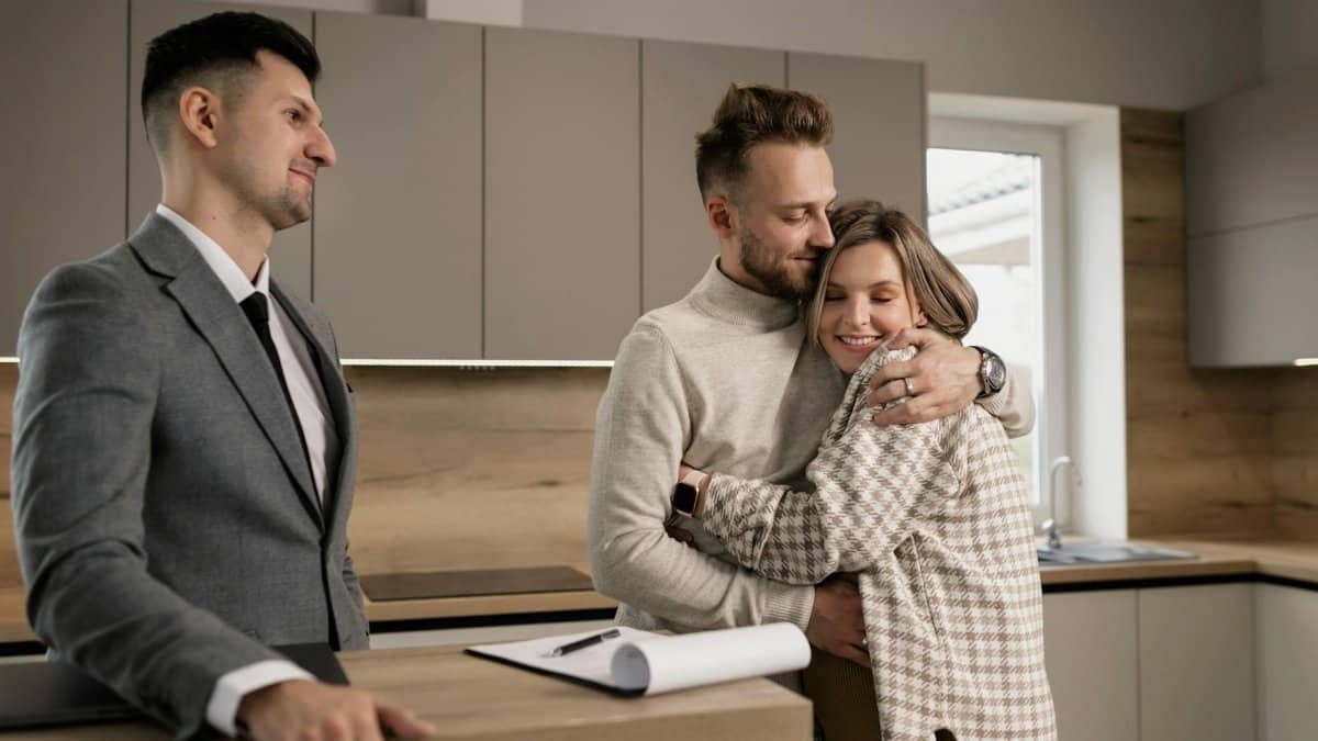 Happy couple embracing in their new home with a real estate agent present.