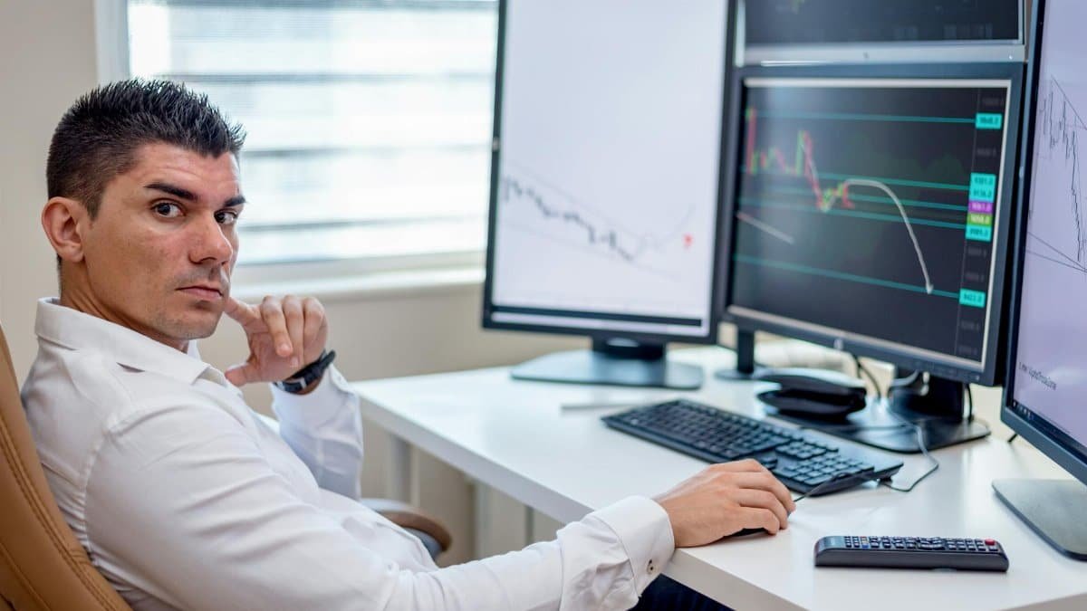 Professional stock market analyst monitoring data on multiple screens at a workstation.